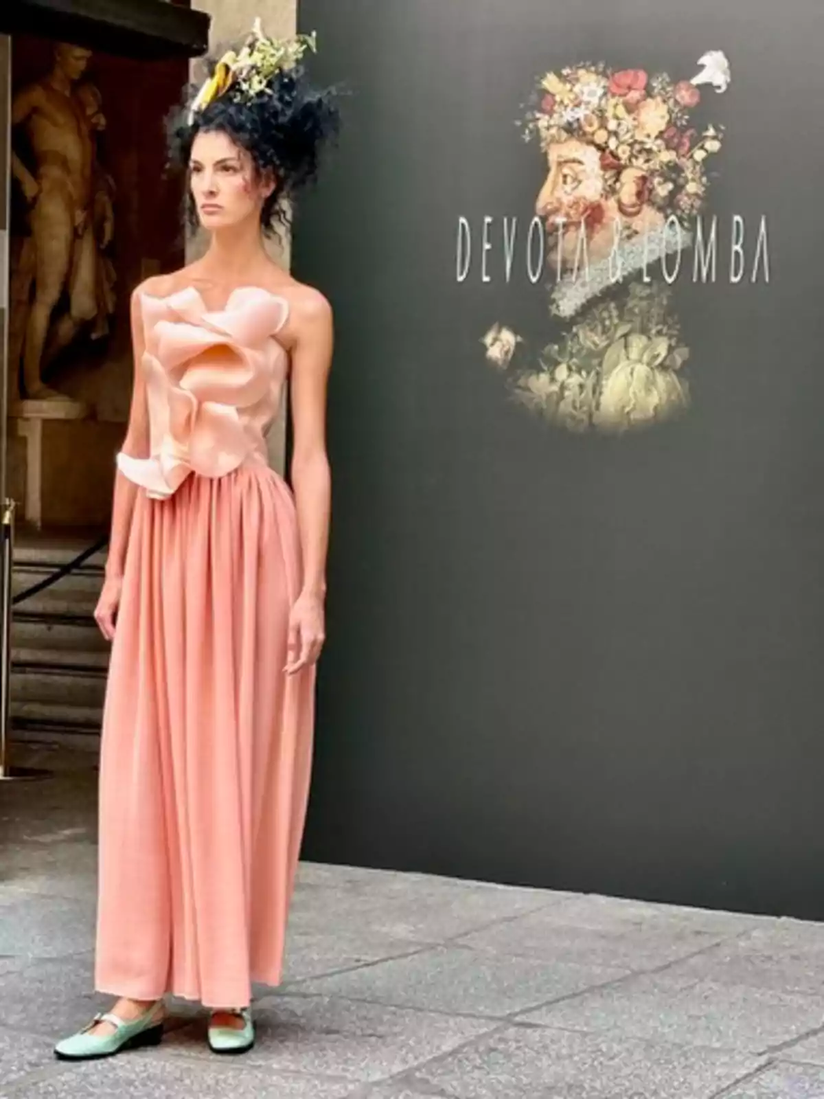 Una modelo con un vestido rosa y un tocado floral posa frente a un cartel de Devota & Lomba.