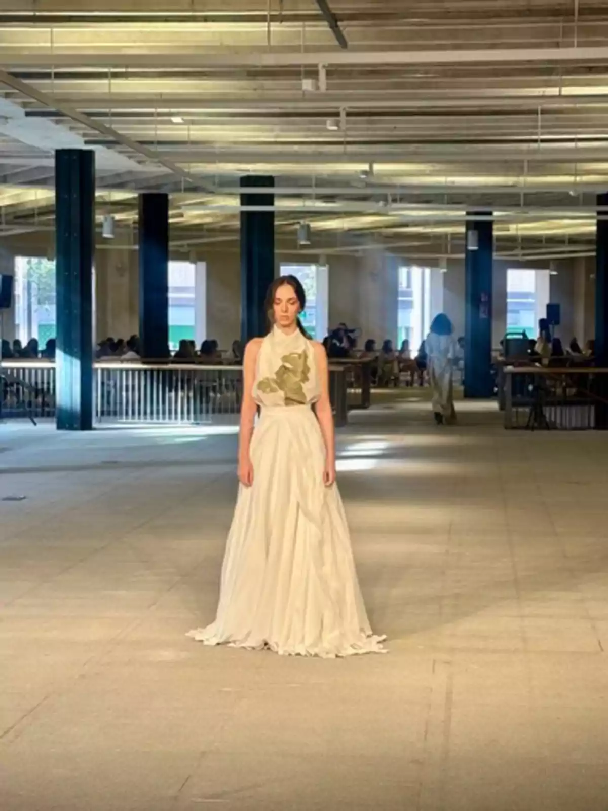 Una modelo con un vestido blanco largo camina por una pasarela en un espacio industrial con techos altos y columnas negras en el desfile de Pilar Dalbat.