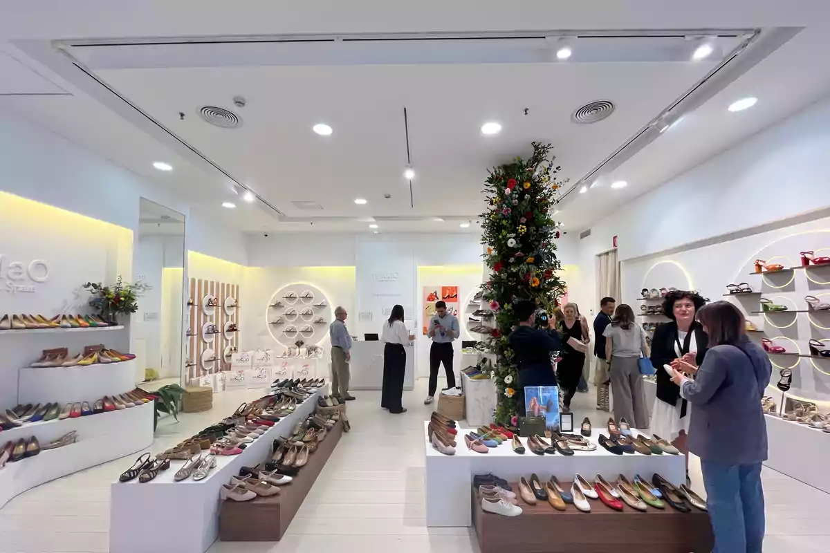 Una tienda de zapatos con estantes llenos de calzado de diferentes colores y estilos, personas conversando y un pilar decorado con flores en el centro.