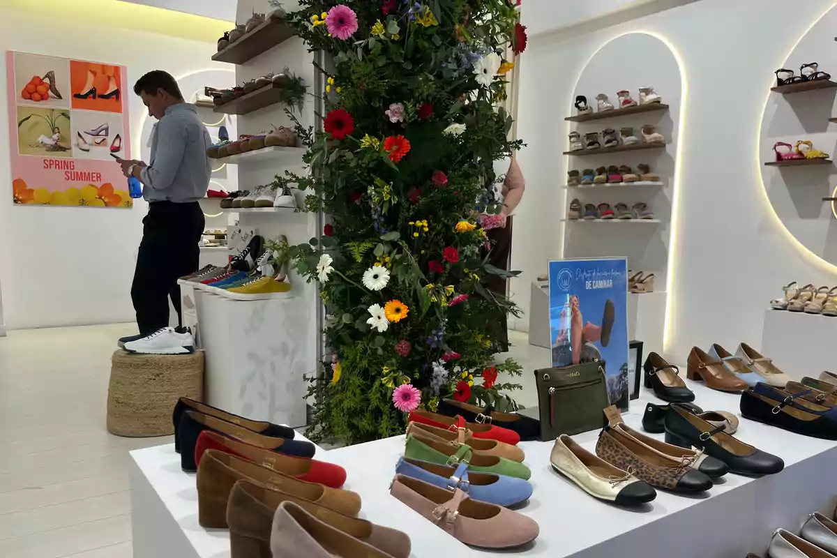 Una tienda de zapatos con una decoración floral en el centro, donde se exhiben diferentes estilos de calzado en estantes y mesas, mientras un hombre revisa su teléfono. Una tienda de zapatos con una decoración floral en el centro, donde se exhiben diferentes estilos de calzado en estantes y mesas, mientras un hombre revisa su teléfono.