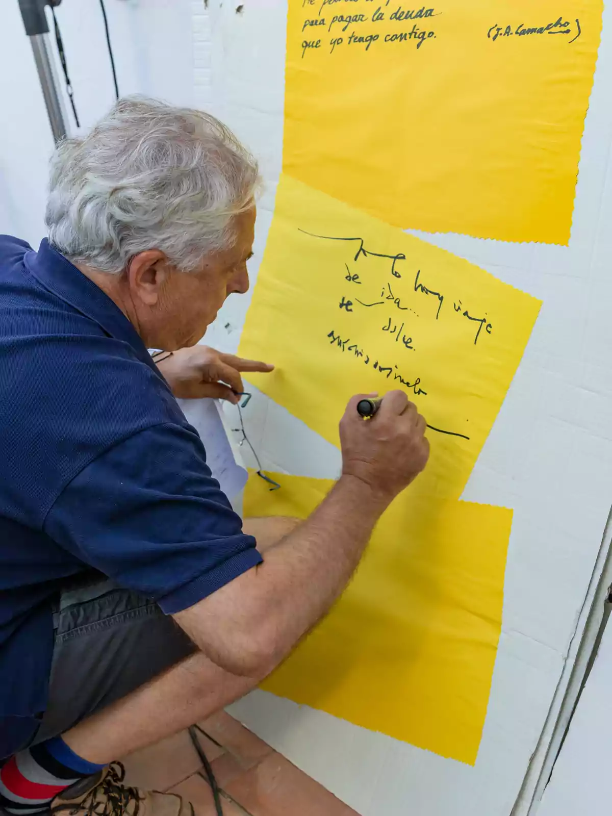 Un hombre mayor escribiendo con un marcador negro sobre un papel amarillo pegado en una pared.