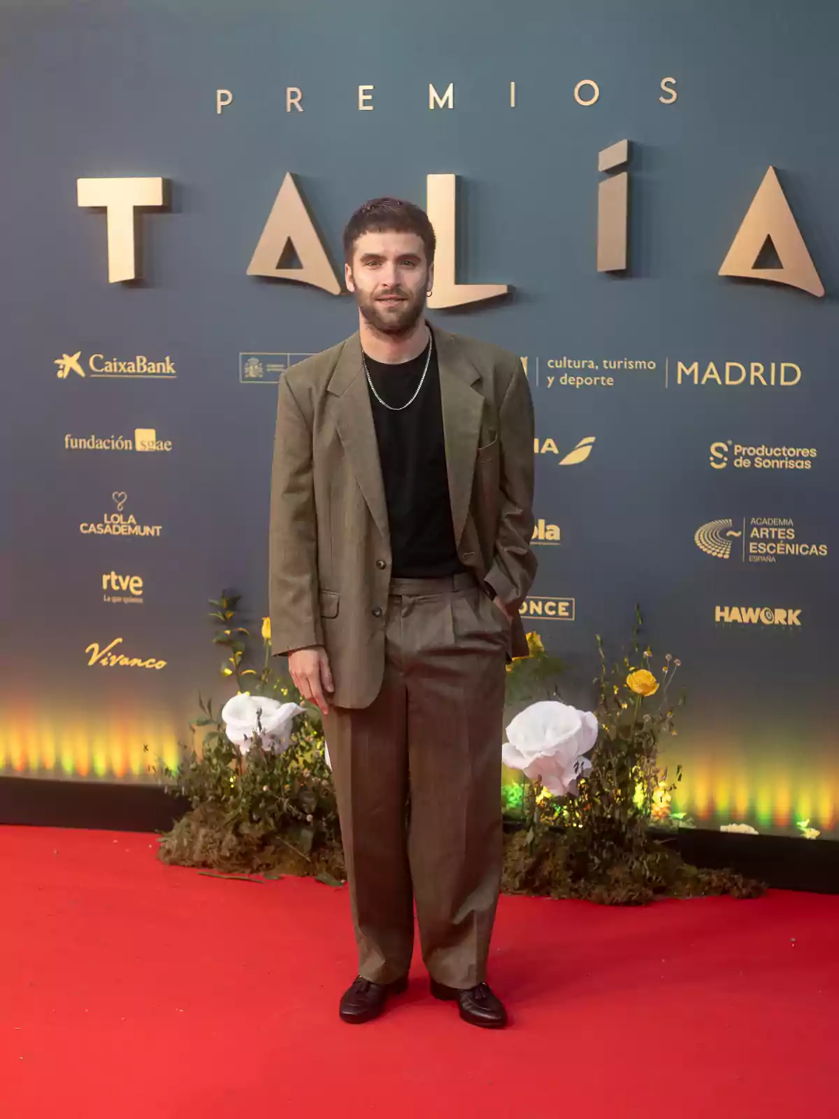 Un hombre con traje marrón y camiseta negra está de pie sobre una alfombra roja frente a un fondo que dice 