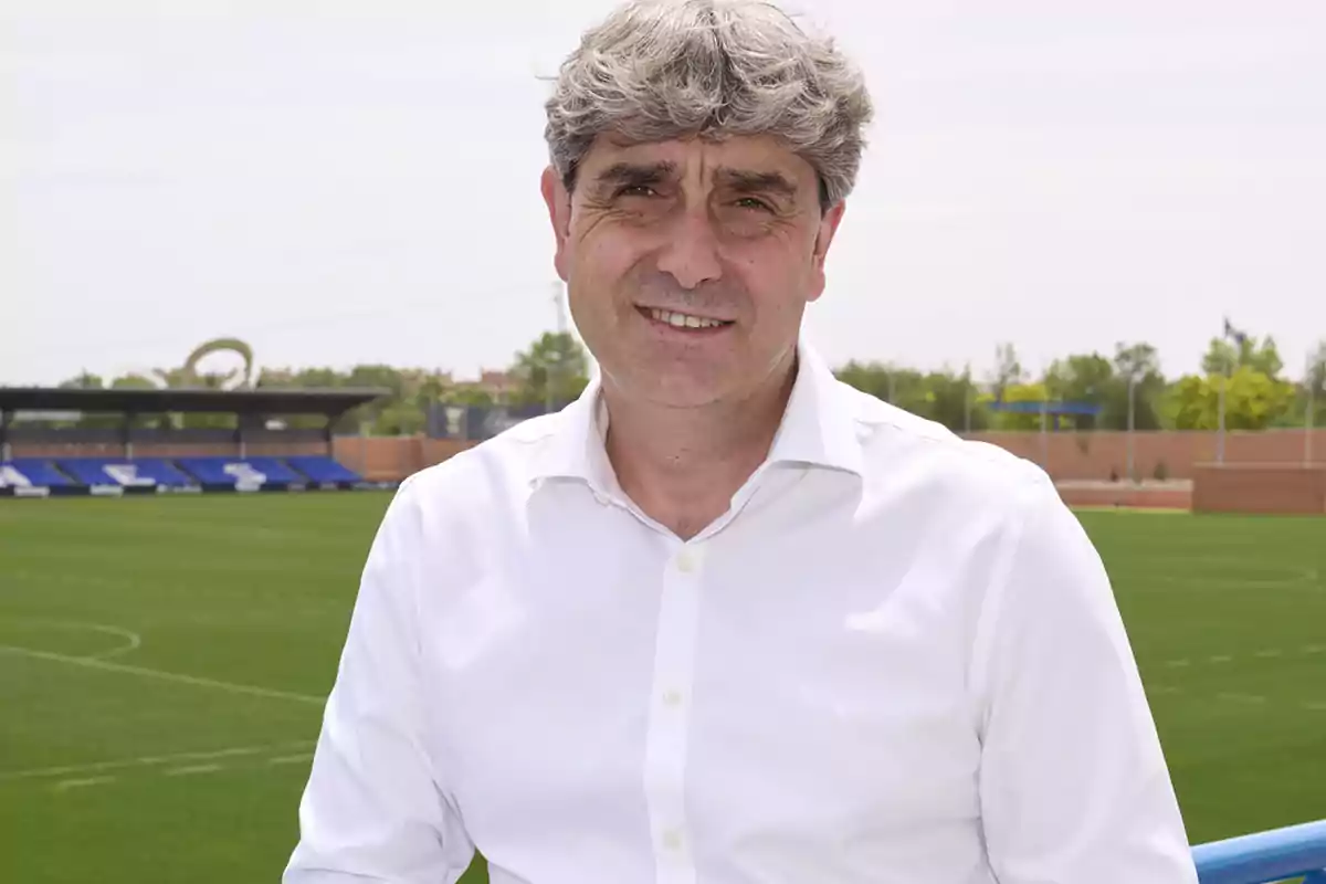 Hombre de cabello canoso y camisa blanca sonriendo frente a un campo de fútbol Hombre de cabello canoso y camisa blanca sonriendo frente a un campo de fútbol