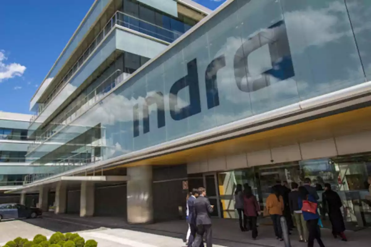 Edificio de oficinas con un gran letrero de "Indra" en la fachada y personas entrando por la puerta principal.