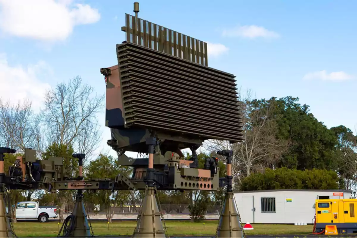 En la imagen se puede observar un radar militar de gran tamaño montado sobre una estructura de soporte, con un fondo de árboles y un cielo parcialmente nublado.