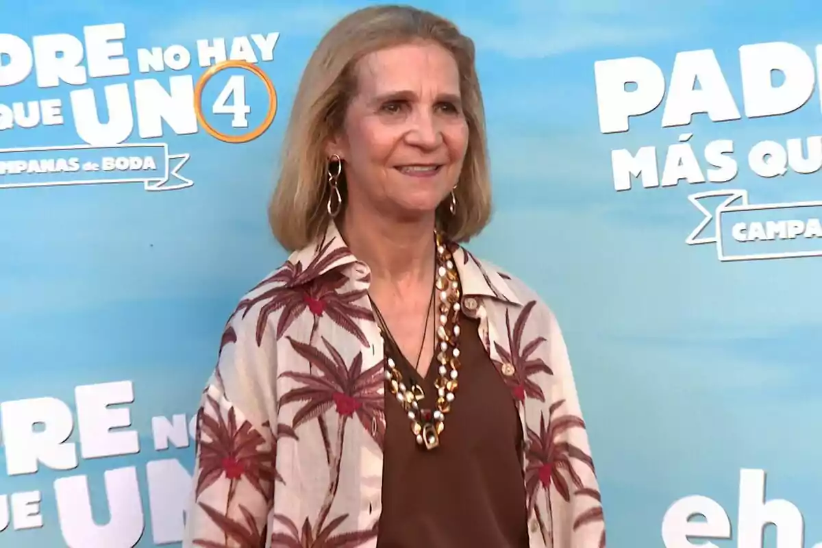 Infanta Elena con una camisa de palmeras y un collar de perlas posando frente a un fondo promocional de una película. Infanta Elena con una camisa de palmeras y un collar de perlas posando frente a un fondo promocional de una película.