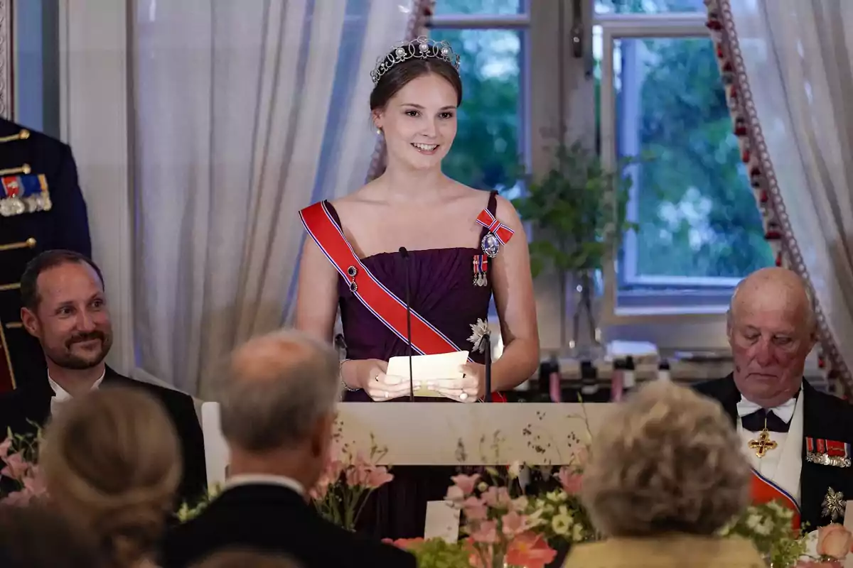 Ingrid Alexandra de Noruega con una tiara y vestido formal da un discurso en un evento elegante, rodeada de personas sentadas en una mesa decorada con flores. Ingrid Alexandra de Noruega con una tiara y vestido formal da un discurso en un evento elegante, rodeada de personas sentadas en una mesa decorada con flores.