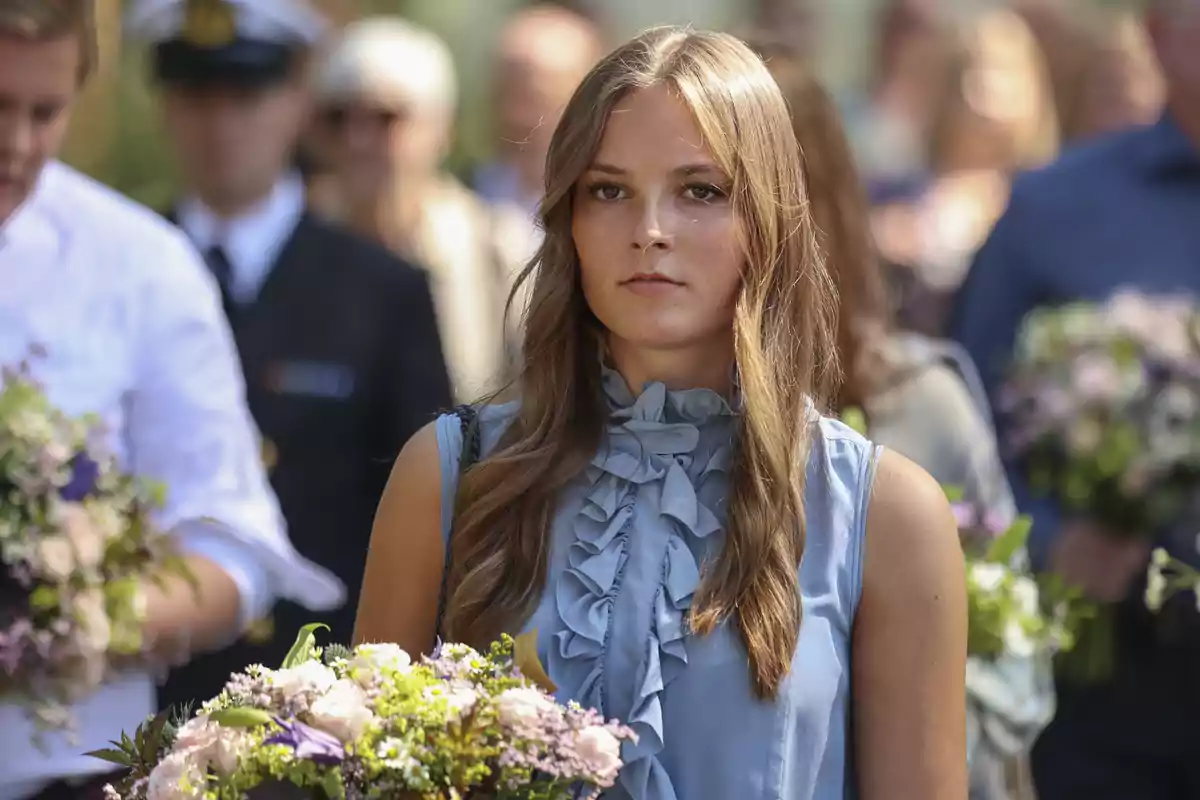 Ingrid Alexandra lleva un vestido azul con volantes mientras sostiene un ramo de flores, rodeada de personas en un evento al aire libre. Ingrid Alexandra lleva un vestido azul con volantes mientras sostiene un ramo de flores, rodeada de personas en un evento al aire libre.