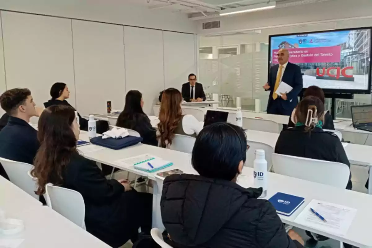 Un grupo de personas en una sala de conferencias escuchando a un presentador frente a una pantalla grande.