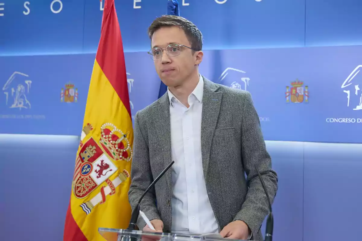 Un hombre con gafas y chaqueta gris está de pie frente a un atril con micrófonos en una sala con la bandera de España y el escudo del Congreso de los Diputados.