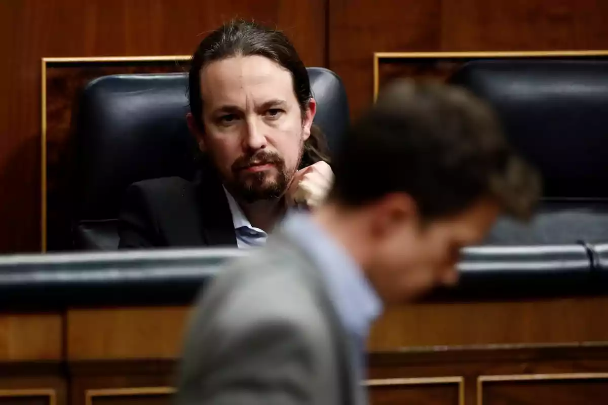 Un hombre con barba y cabello largo está sentado en un asiento de cuero oscuro en un entorno formal, mientras otra persona aparece desenfocada en primer plano.