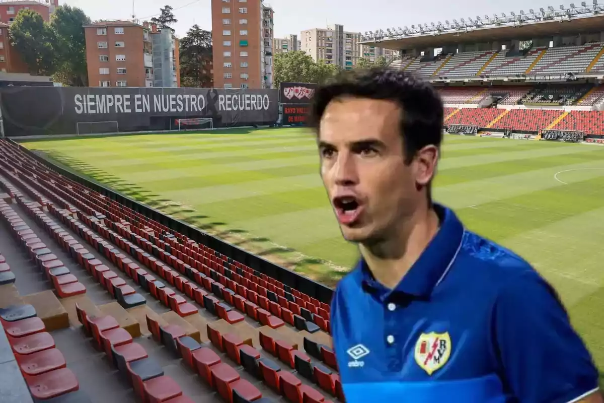 Un hombre con una camiseta azul del Rayo Vallecano aparece en primer plano frente a un estadio de fútbol vacío con gradas rojas y negras y un cartel que dice "Siempre en nuestro recuerdo".