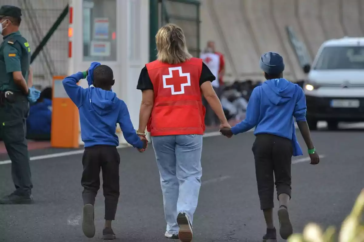 Una trabajadora de la Cruz Roja camina de la mano con dos niños mientras un guardia civil observa.