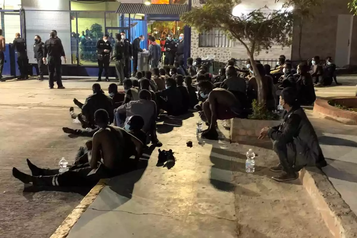 Un grupo de personas sentadas en el suelo al aire libre, con varias botellas de agua a su alrededor, mientras algunos oficiales de seguridad están de pie cerca de una entrada cercada.