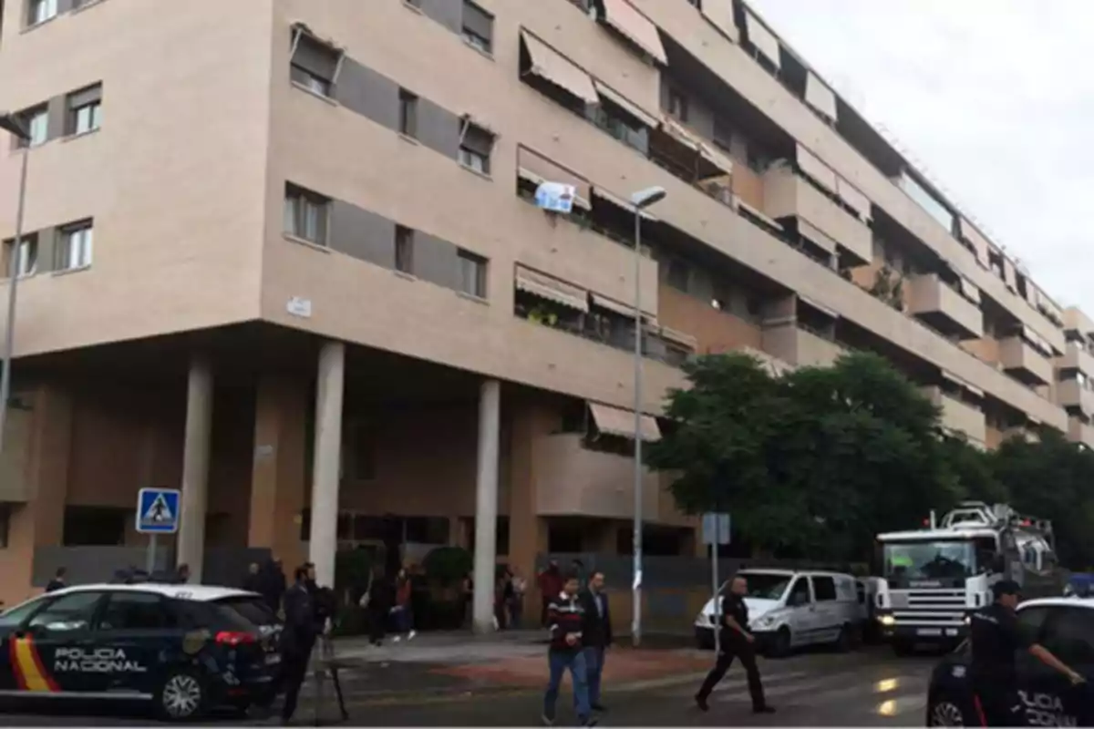 Edificio de apartamentos con vehículos de la Policía Nacional estacionados en la calle y varias personas caminando por la acera.