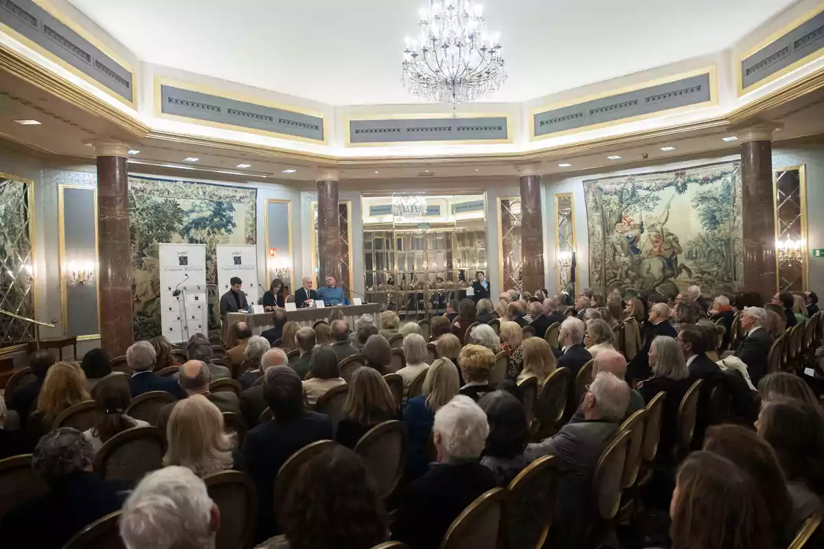 Una conferencia en un elegante salón con un público numeroso y un panel de oradores al frente.