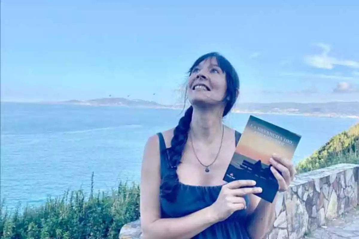 Mujer sonriendo y sosteniendo un libro con el mar y un paisaje costero de fondo.