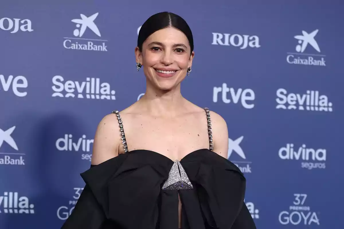 Una persona con un vestido negro elegante posando frente a un fondo azul con logotipos de patrocinadores.