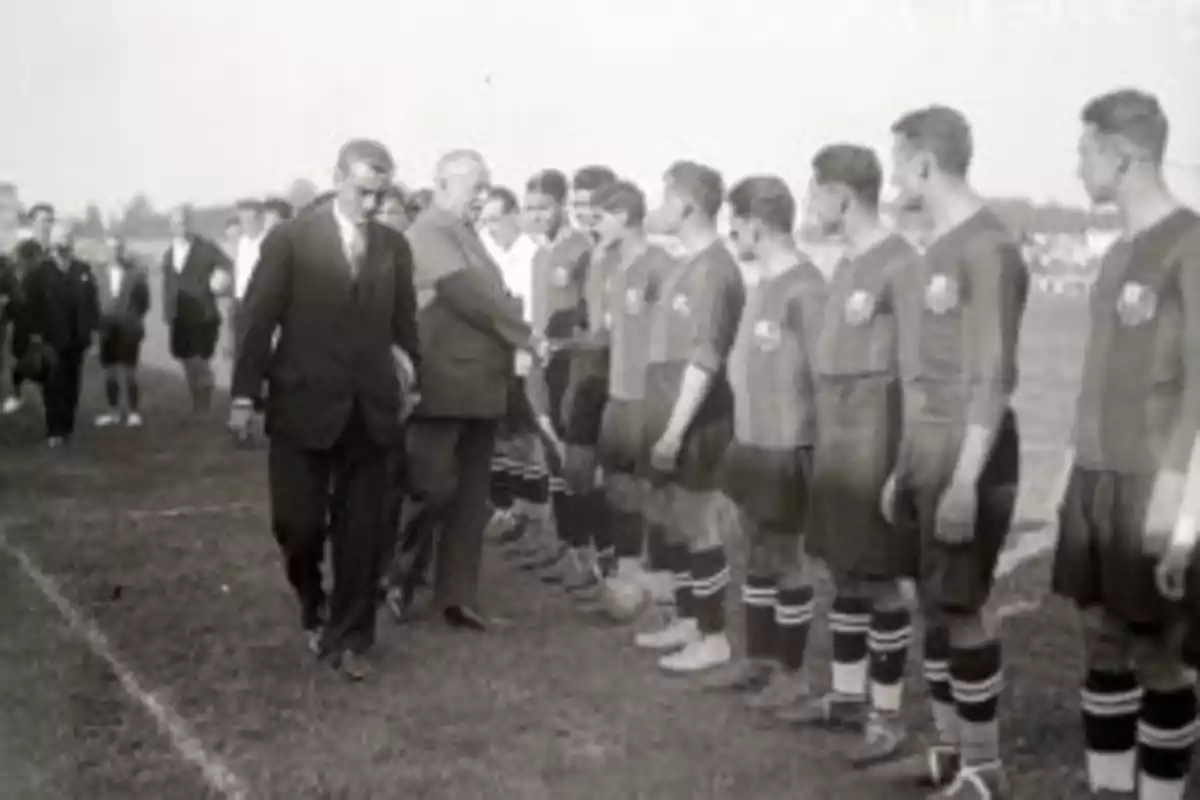 Un grupo de jugadores de fútbol alineados en el campo mientras dos hombres de traje los saludan.