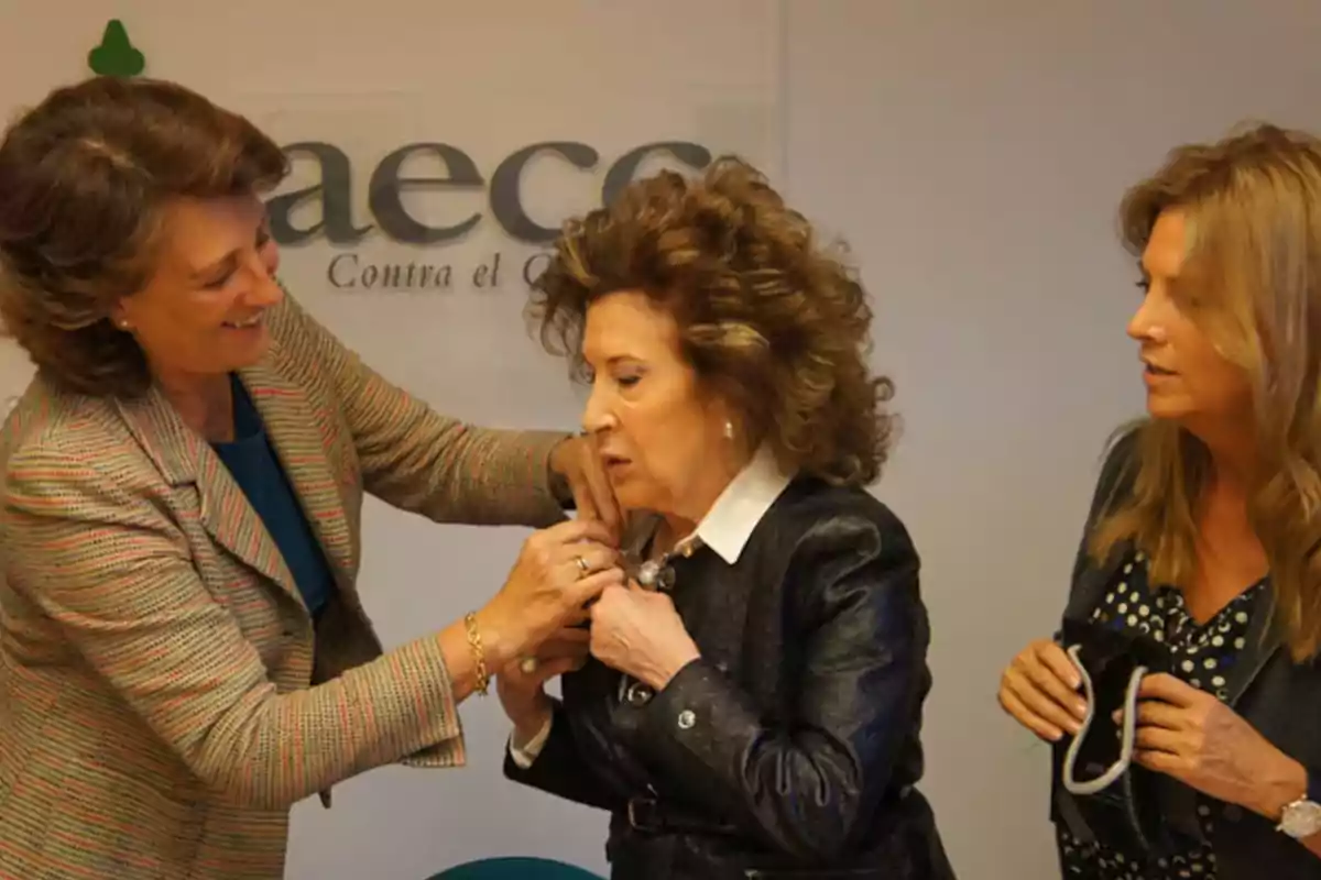 Tres mujeres interactúan en un evento de la aecc mientras una de ellas coloca un pin en la solapa de otra Tres mujeres interactúan en un evento de la aecc mientras una de ellas coloca un pin en la solapa de otra