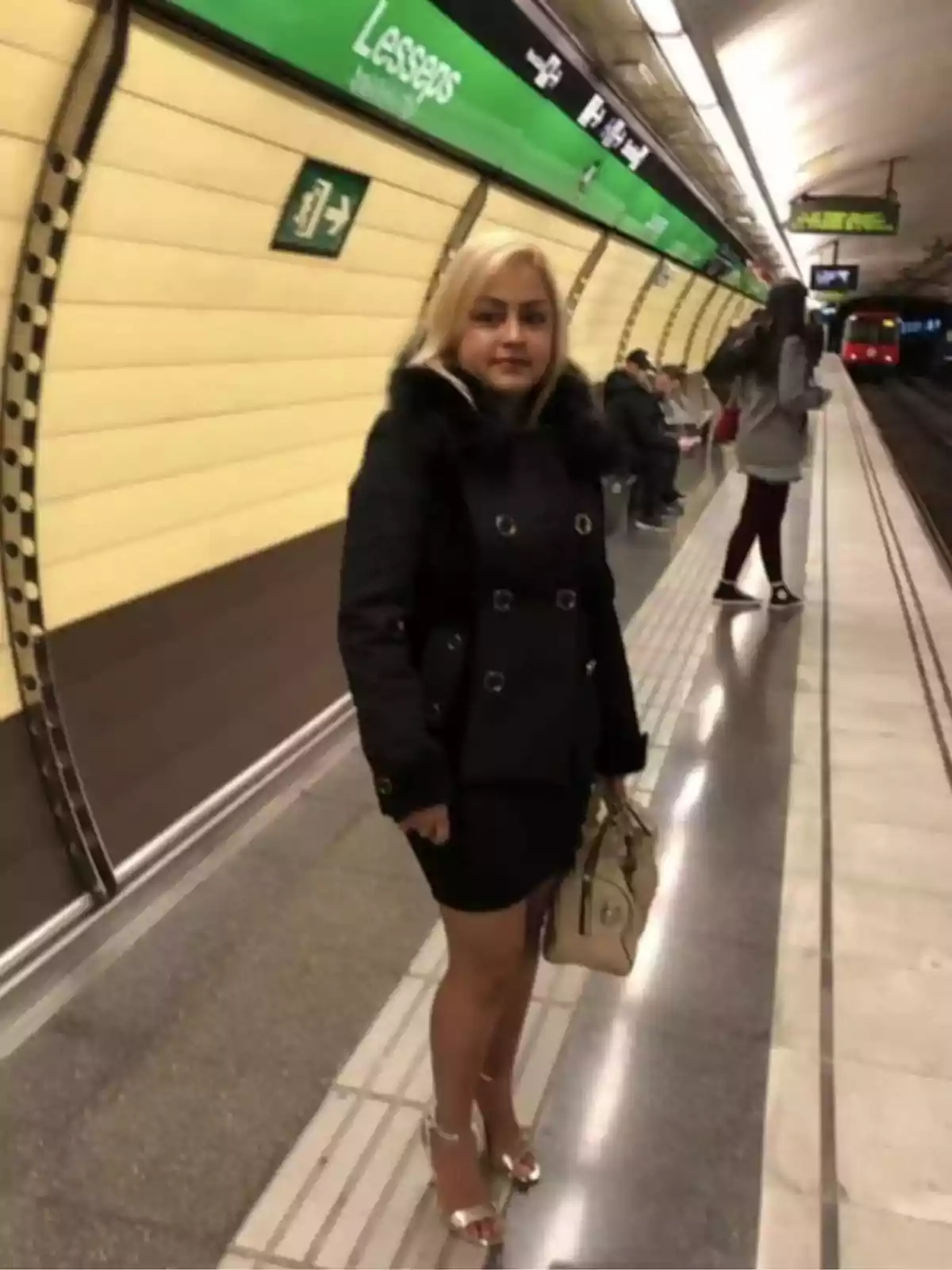 Una mujer rubia con abrigo negro y bolso beige está de pie en una estación de metro llamada Lesseps.