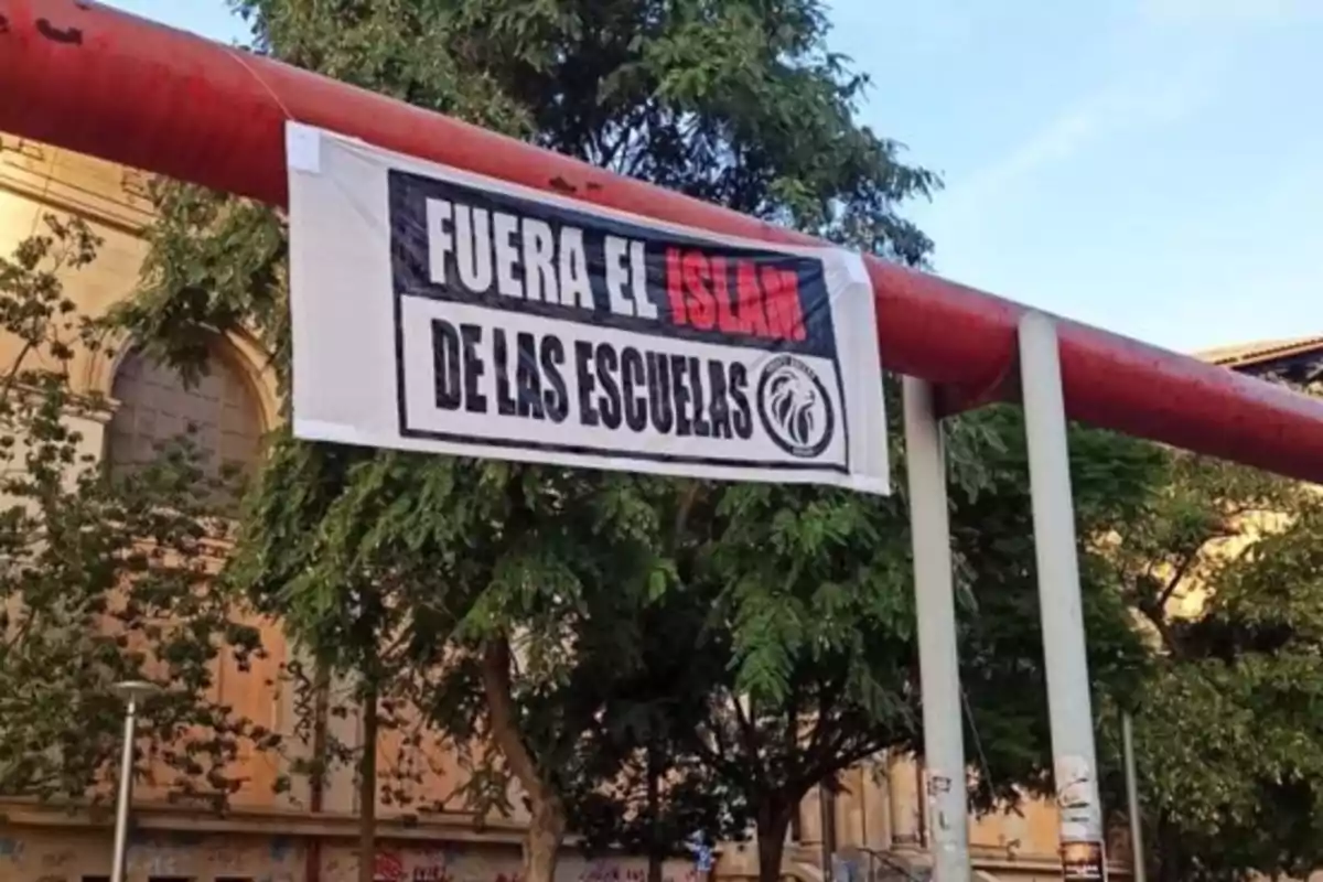 Una pancarta colgada en una estructura metálica roja con un mensaje en contra del islam en las escuelas, rodeada de árboles y un edificio al fondo. Una pancarta colgada en una estructura metálica roja con un mensaje en contra del islam en las escuelas, rodeada de árboles y un edificio al fondo.