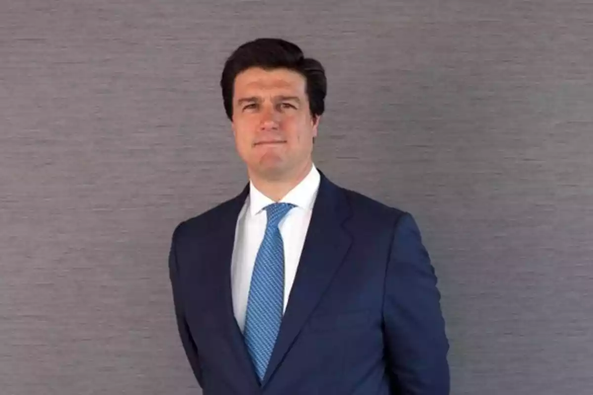 Hombre de cabello oscuro vistiendo traje azul y corbata azul claro posando frente a un fondo gris Hombre de cabello oscuro vistiendo traje azul y corbata azul claro posando frente a un fondo gris
