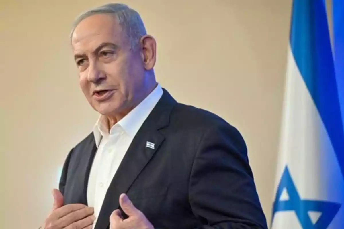 Hombre de cabello canoso con traje oscuro y camisa blanca hablando frente a una bandera de Israel Hombre de cabello canoso con traje oscuro y camisa blanca hablando frente a una bandera de Israel