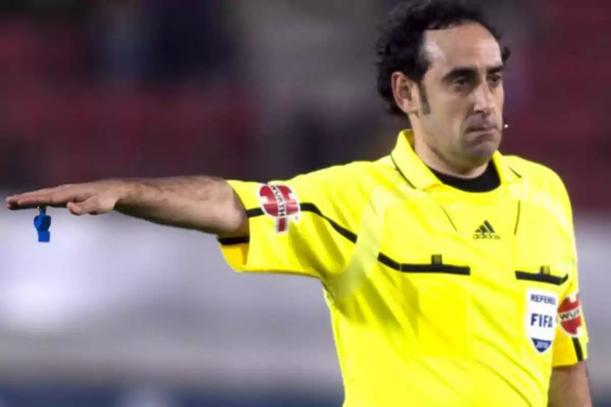 Un árbitro de fútbol con uniforme amarillo señalando con el brazo extendido y sosteniendo un silbato azul.