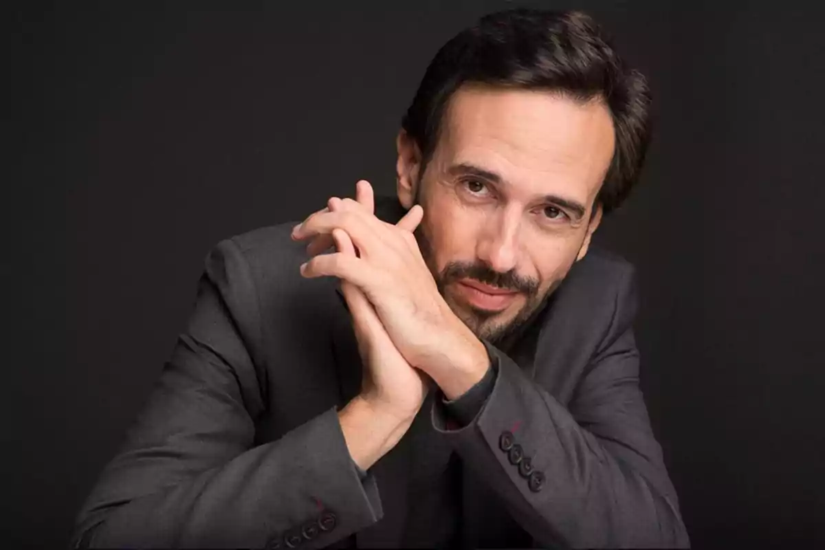 Hombre con barba y cabello oscuro en traje gris posando con las manos entrelazadas sobre un fondo negro. Hombre con barba y cabello oscuro en traje gris posando con las manos entrelazadas sobre un fondo negro.