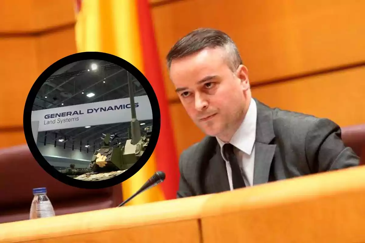 Un hombre en traje sentado en una mesa con un micrófono, junto a un círculo que muestra un tanque y un cartel de General Dynamics Land Systems.