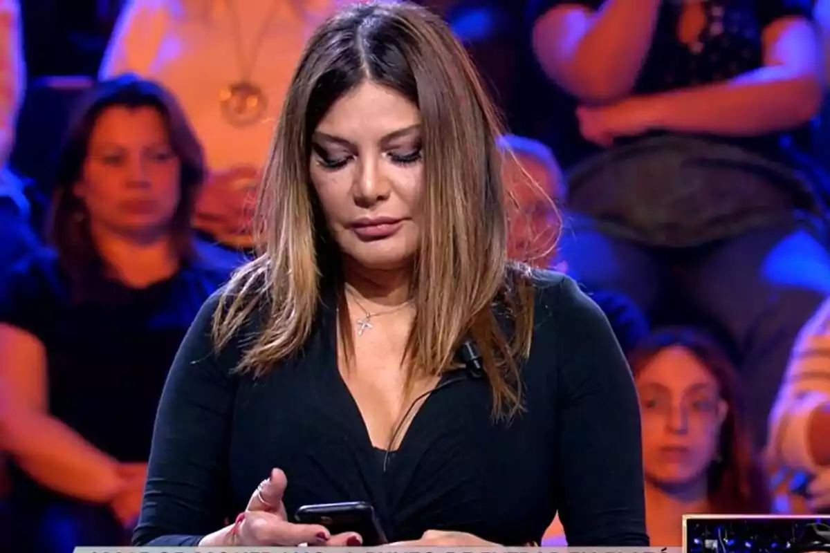 Ivonne Reyes con cabello castaño y un vestido negro está mirando su teléfono en un entorno con público al fondo en 'De Viernes'. Ivonne Reyes con cabello castaño y un vestido negro está mirando su teléfono en un entorno con público al fondo en 'De Viernes'.