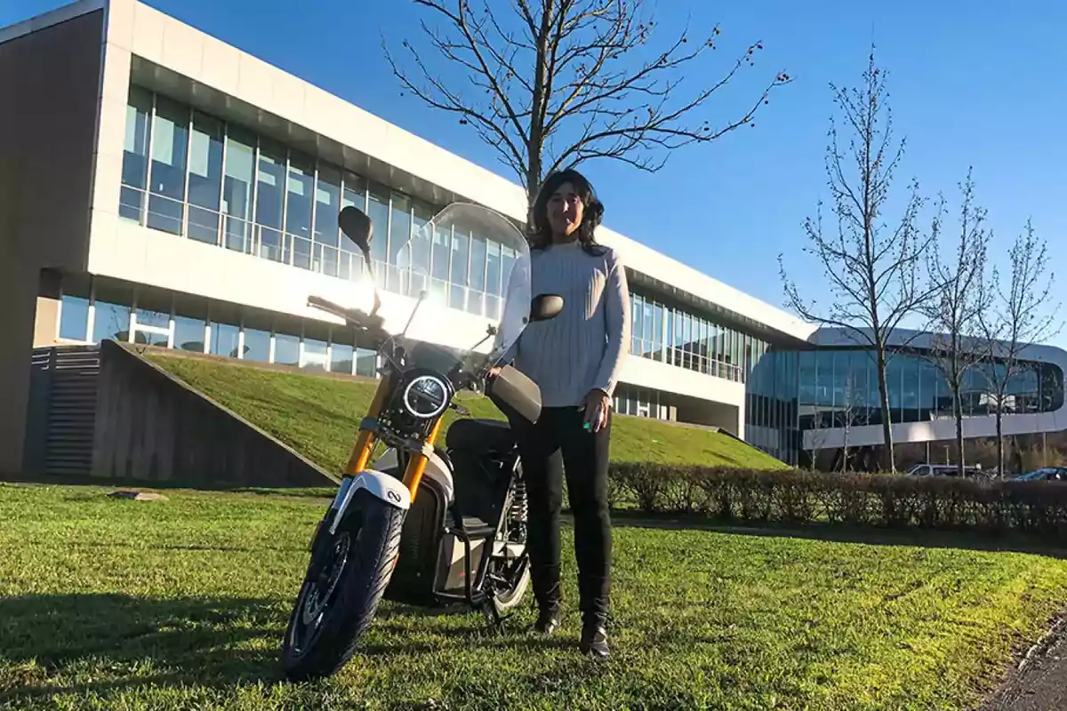 Una mujer de pie junto a una motocicleta en un área verde frente a un edificio moderno.