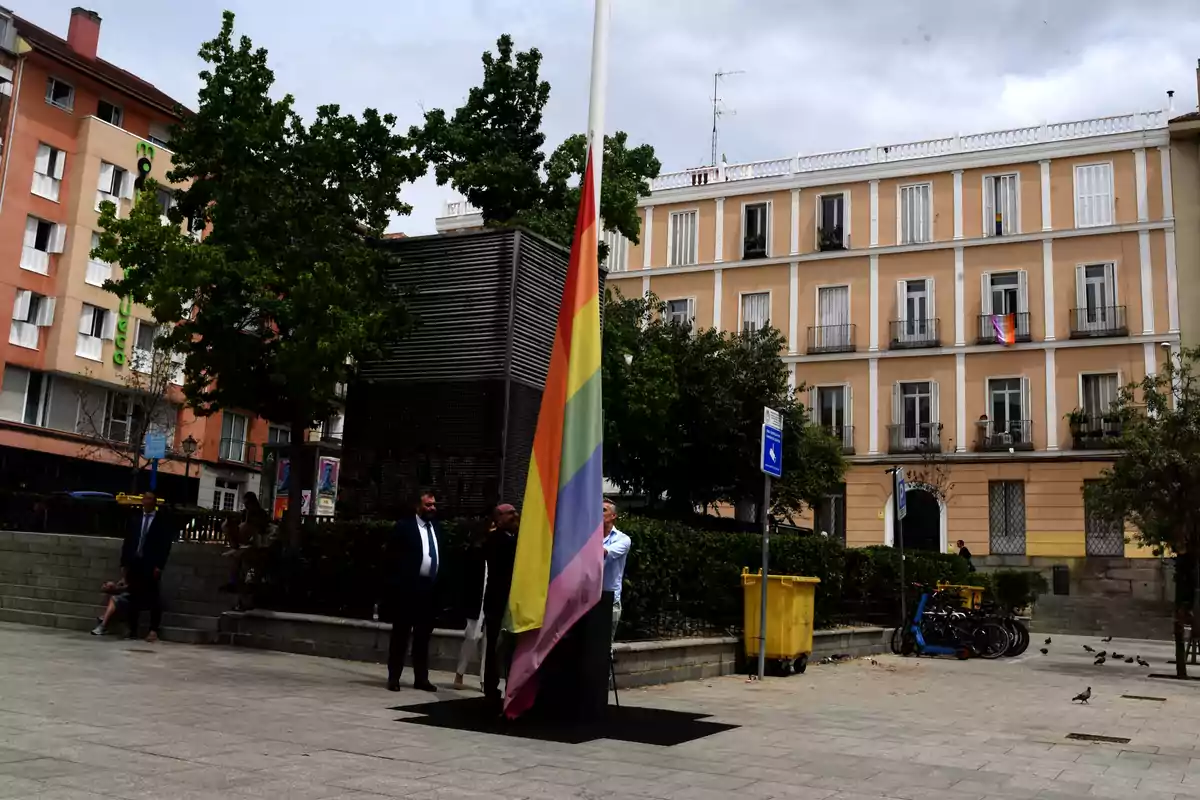 Personas izando una bandera arcoíris en una plaza urbana con edificios residenciales de fondo.