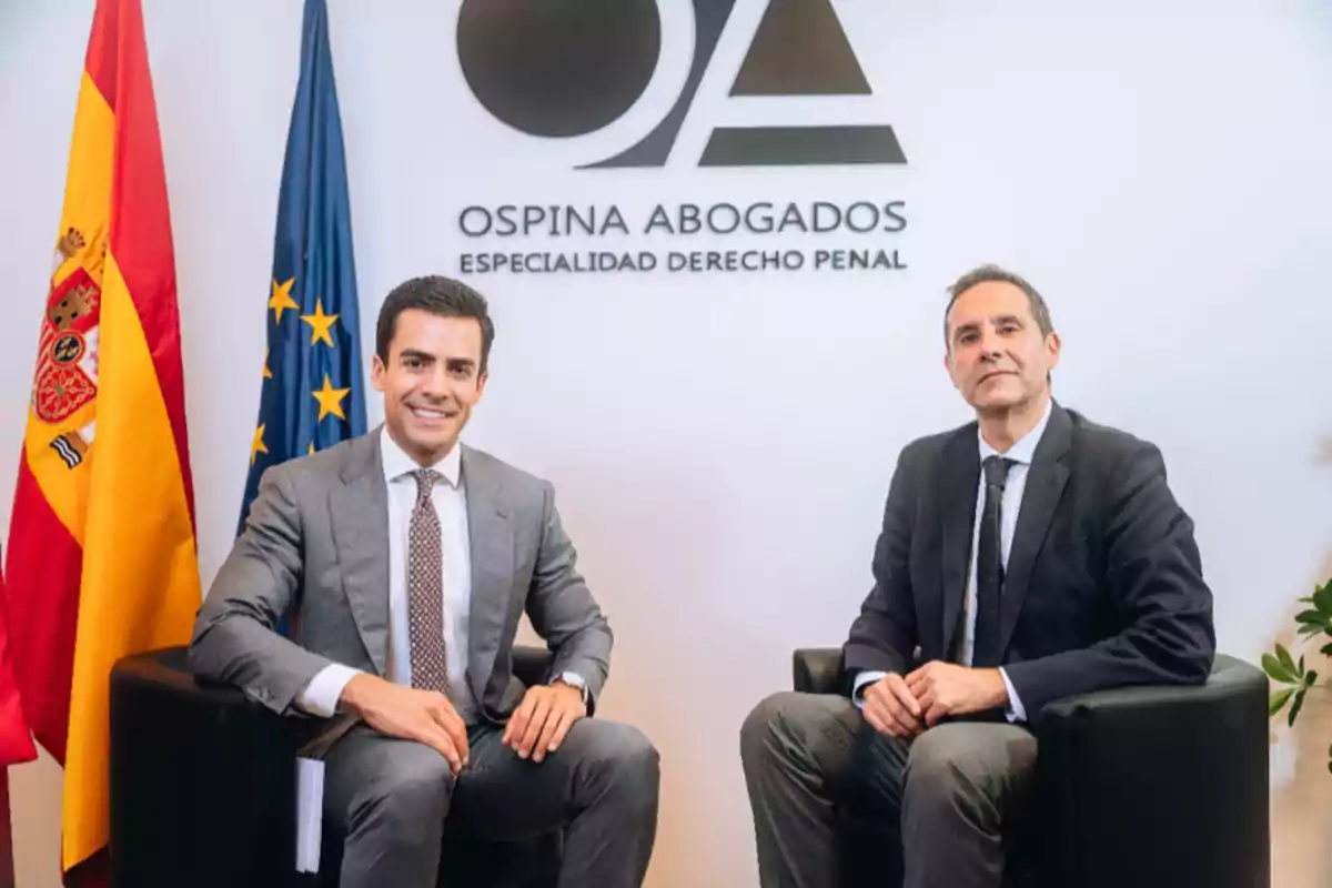 Dos hombres en trajes sentados en una oficina de abogados con banderas de España y la Unión Europea en el fondo.
