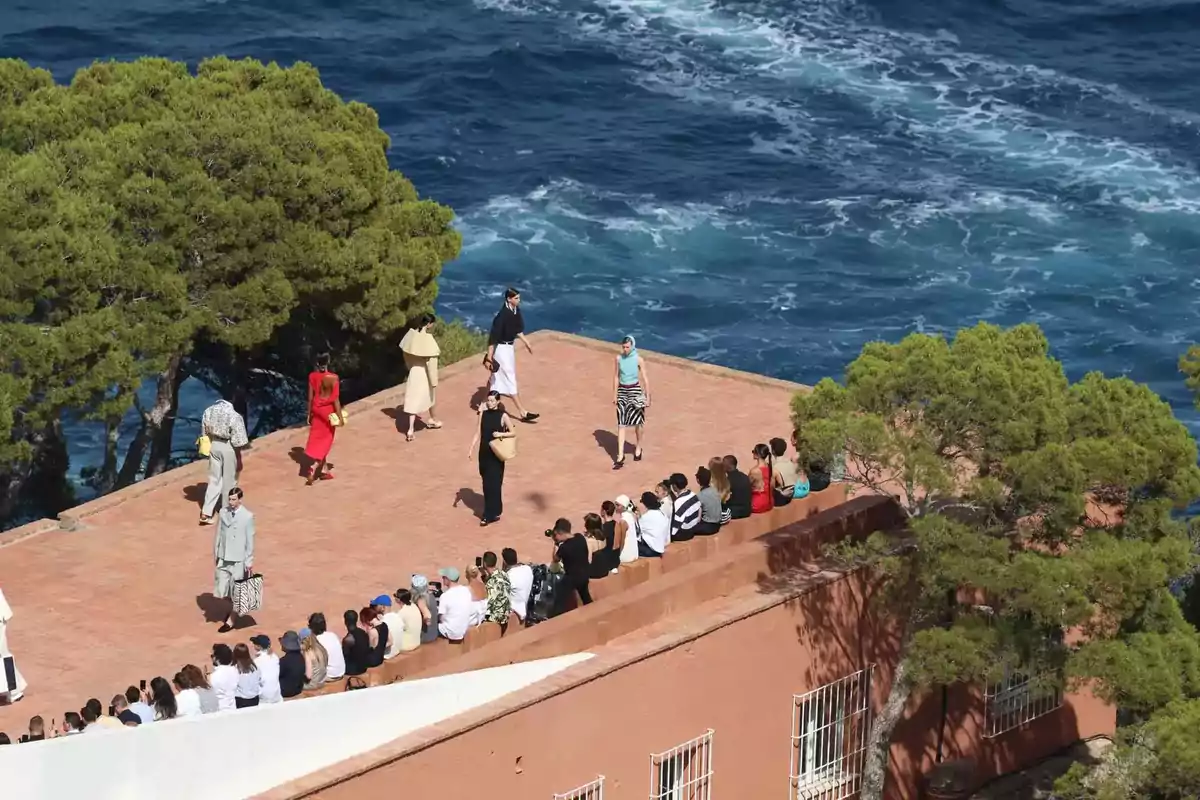 Desfile de moda al aire libre con modelos caminando sobre una plataforma elevada junto al mar y espectadores sentados observando.