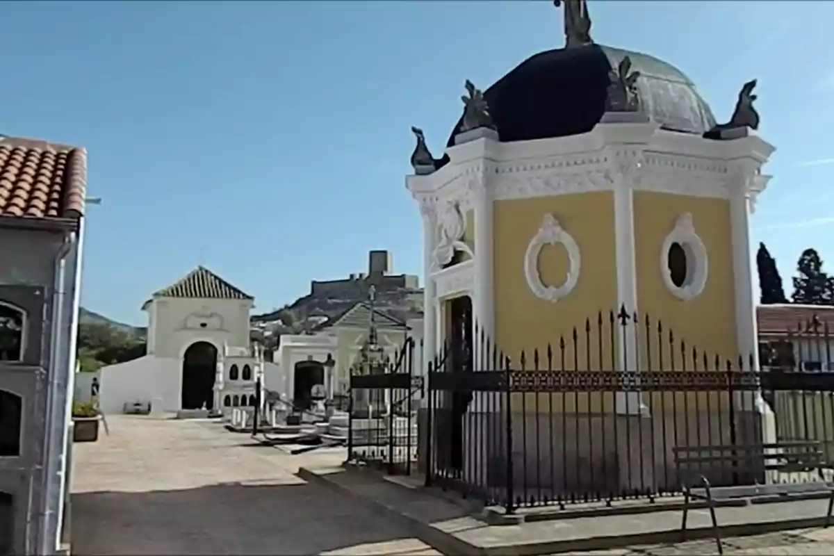 Vista de un cementerio con una capilla amarilla y una estructura blanca al fondo bajo un cielo despejado.