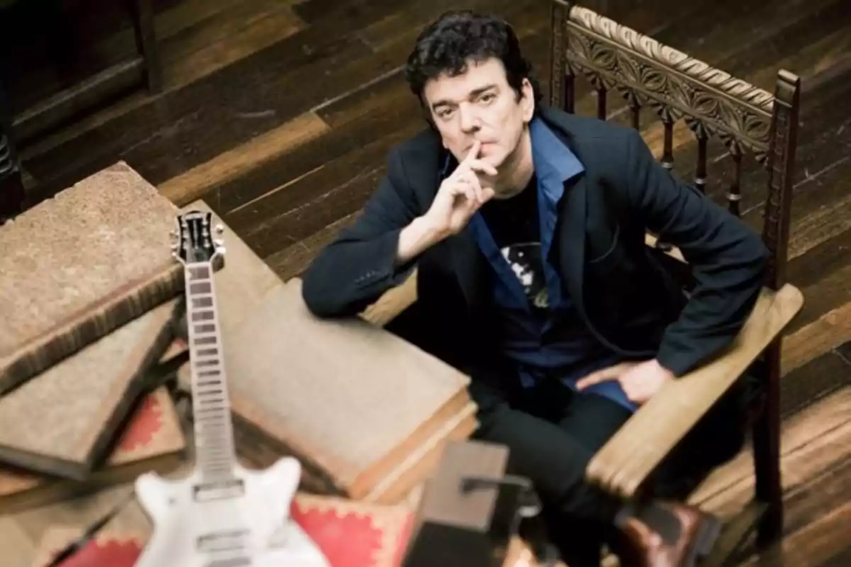 Hombre sentado en una silla de madera junto a una guitarra y varios libros grandes sobre una mesa.