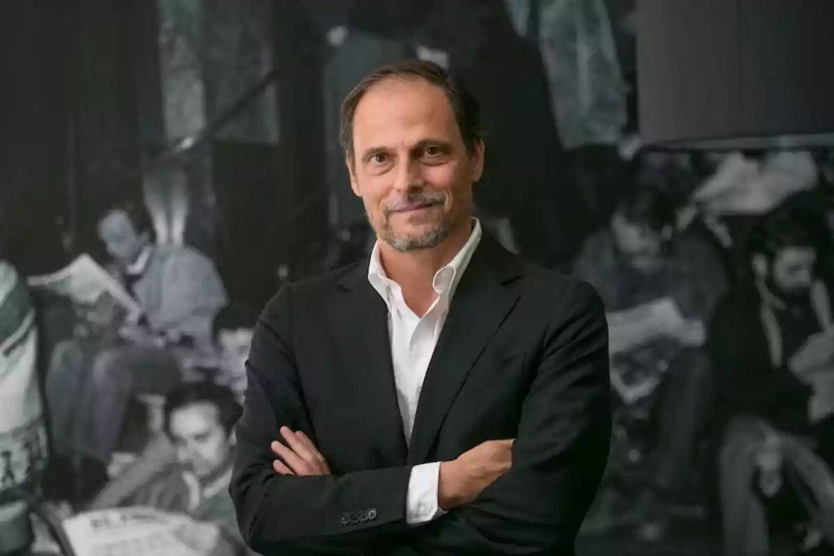 Hombre de mediana edad con barba y cabello corto, vestido con traje oscuro y camisa blanca, posando con los brazos cruzados frente a un fondo en blanco y negro de personas leyendo. Hombre de mediana edad con barba y cabello corto, vestido con traje oscuro y camisa blanca, posando con los brazos cruzados frente a un fondo en blanco y negro de personas leyendo.