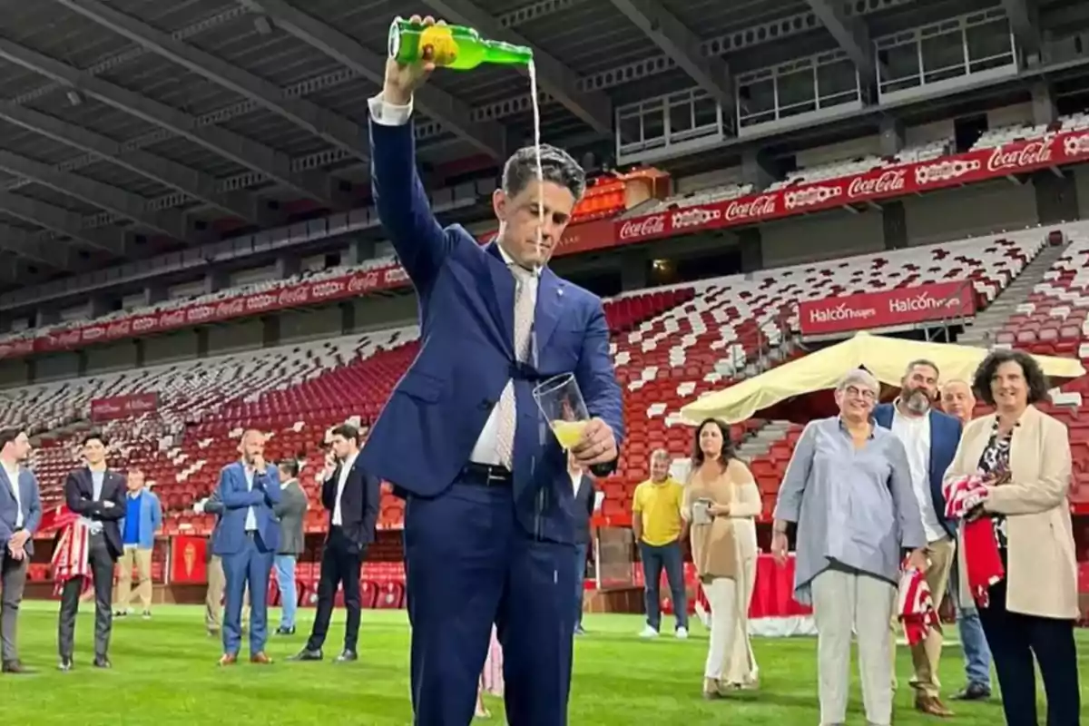 Un hombre en traje está sirviendo sidra desde una botella verde en un vaso, en un estadio con personas observando. Un hombre en traje está sirviendo sidra desde una botella verde en un vaso, en un estadio con personas observando.