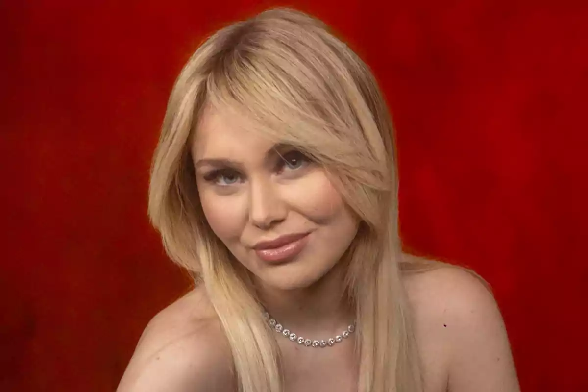 Una mujer rubia con cabello largo y liso, usando un collar de perlas, sobre un fondo rojo.