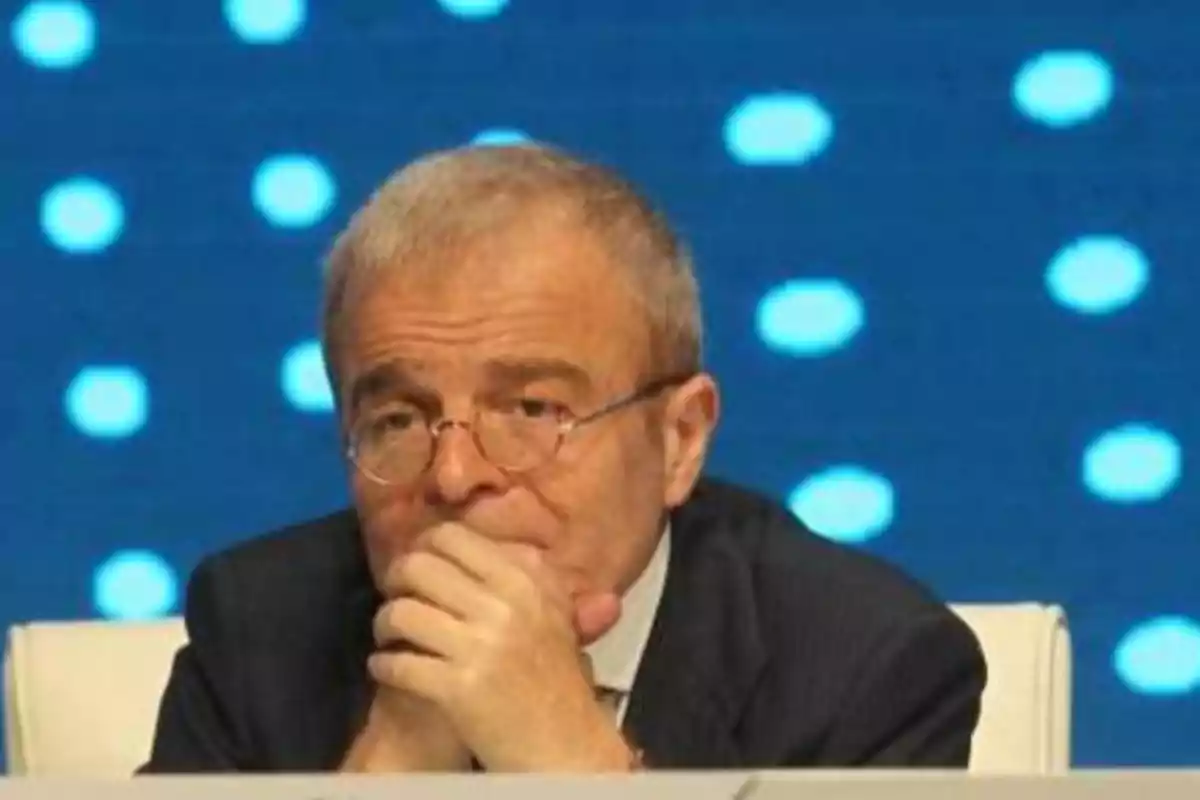 Un hombre con gafas y traje oscuro está sentado con las manos juntas frente a su rostro, sobre un fondo azul con puntos blancos.