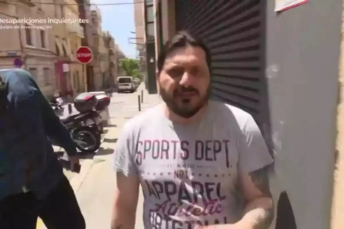 Un hombre con camiseta gris camina por una calle urbana con edificios y una señal de stop al fondo.