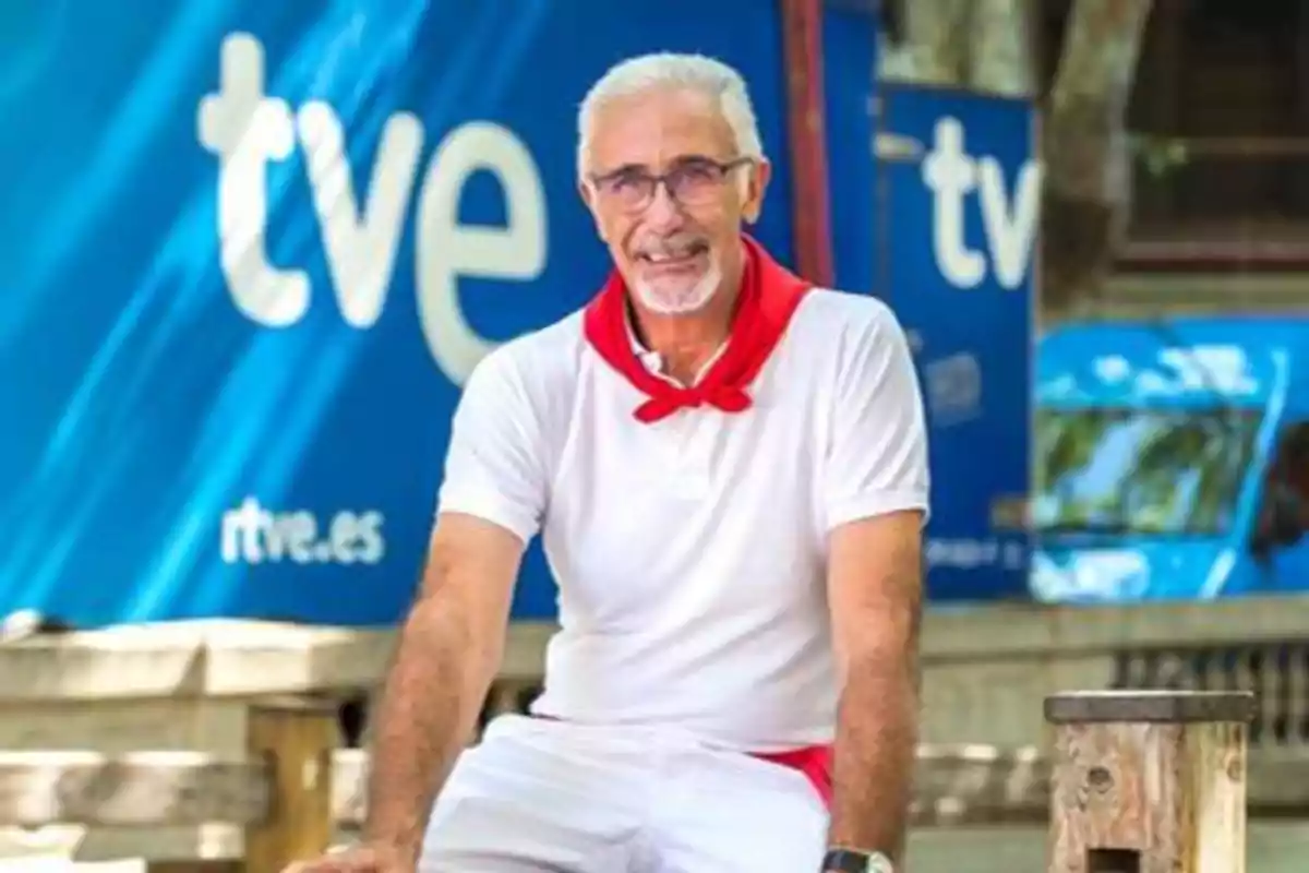 Un hombre mayor con cabello canoso y gafas, vestido con una camiseta blanca y un pañuelo rojo, está sentado frente a un fondo azul con el logotipo de "tve".