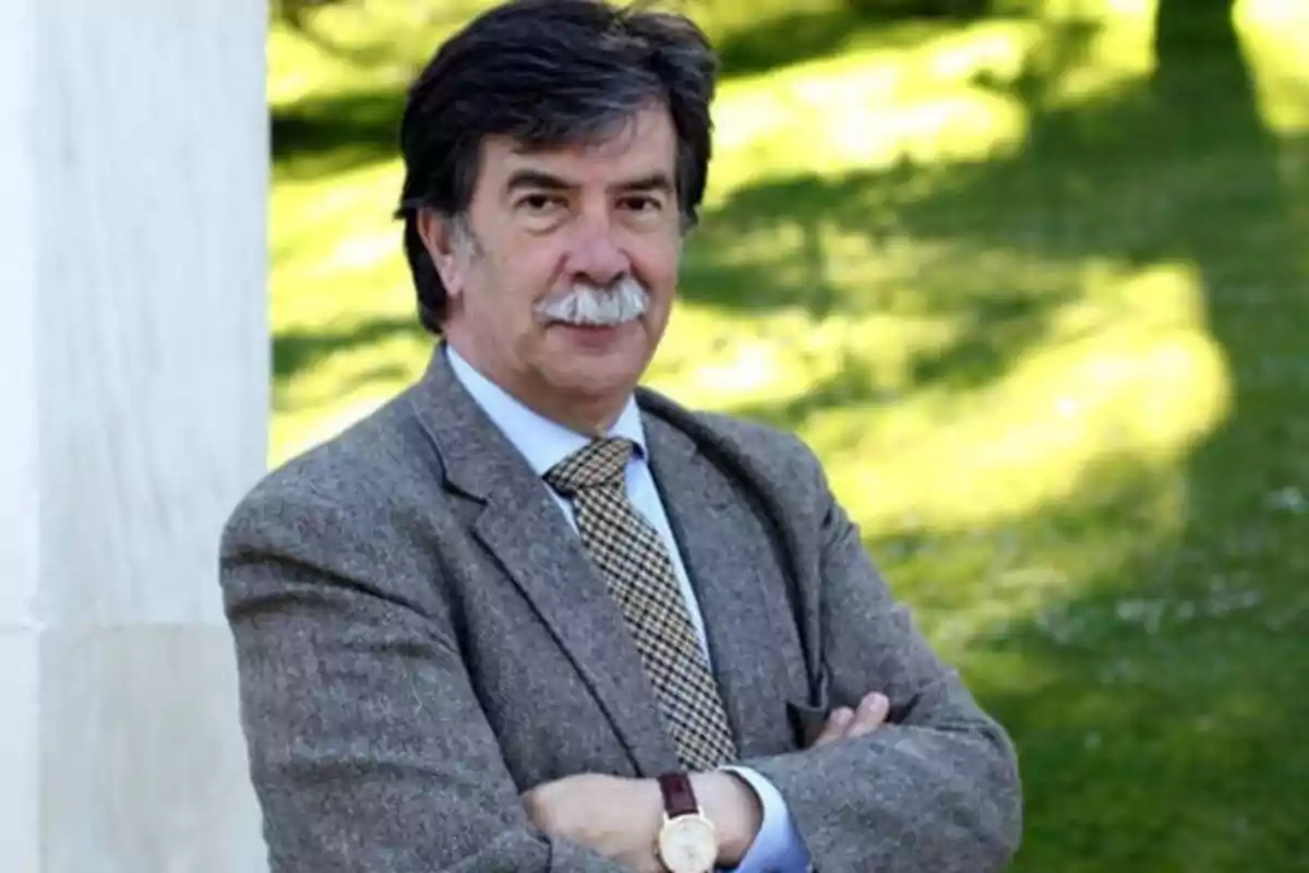 Hombre con bigote y cabello oscuro, vestido con traje gris y corbata, posando con los brazos cruzados en un entorno al aire libre con césped verde.