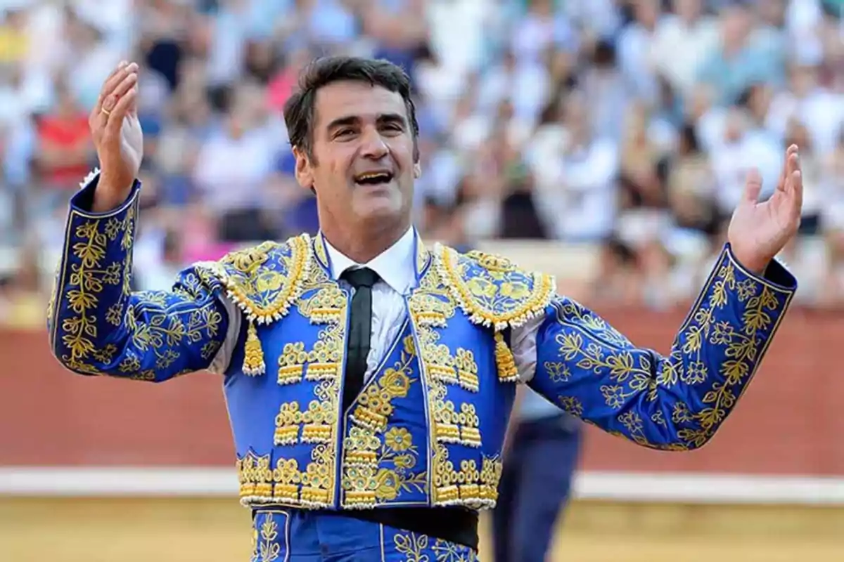 Un torero con traje de luces azul y dorado en una plaza de toros, con los brazos levantados y una expresión de alegría.