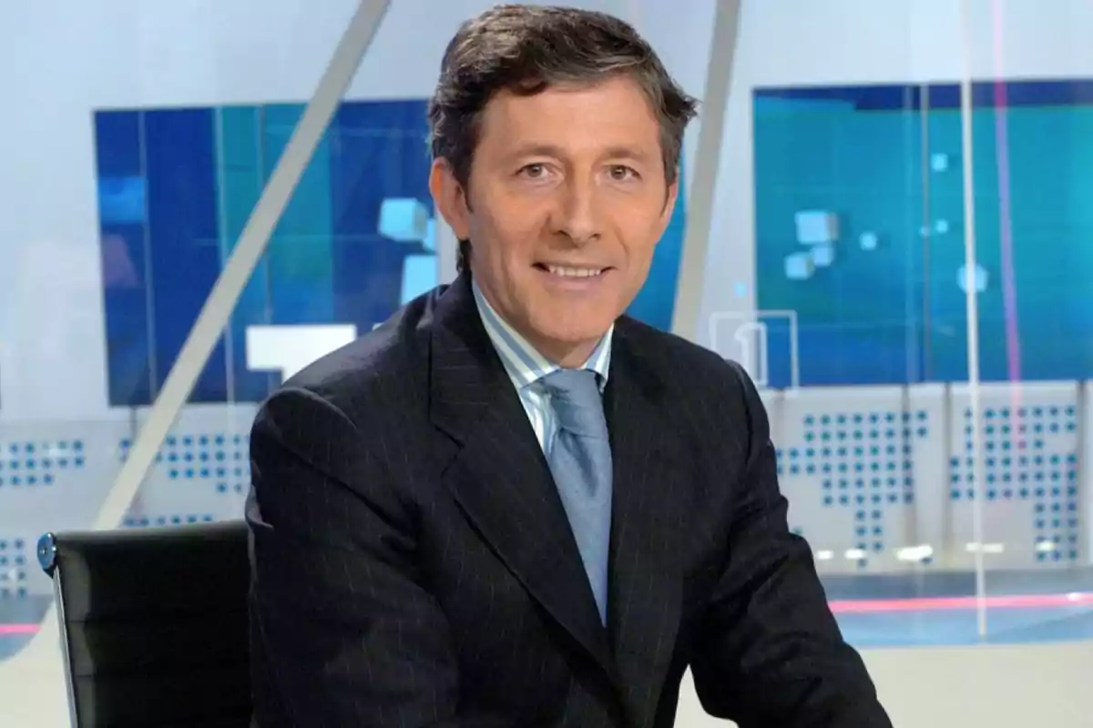Hombre de cabello corto y canoso con traje oscuro y corbata azul sentado en un estudio de televisión moderno Hombre de cabello corto y canoso con traje oscuro y corbata azul sentado en un estudio de televisión moderno