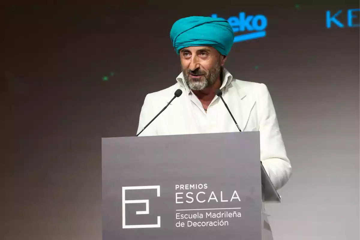 Hombre con turbante azul y traje blanco hablando en un podio durante los Premios Escala de la Escuela Madrileña de Decoración.