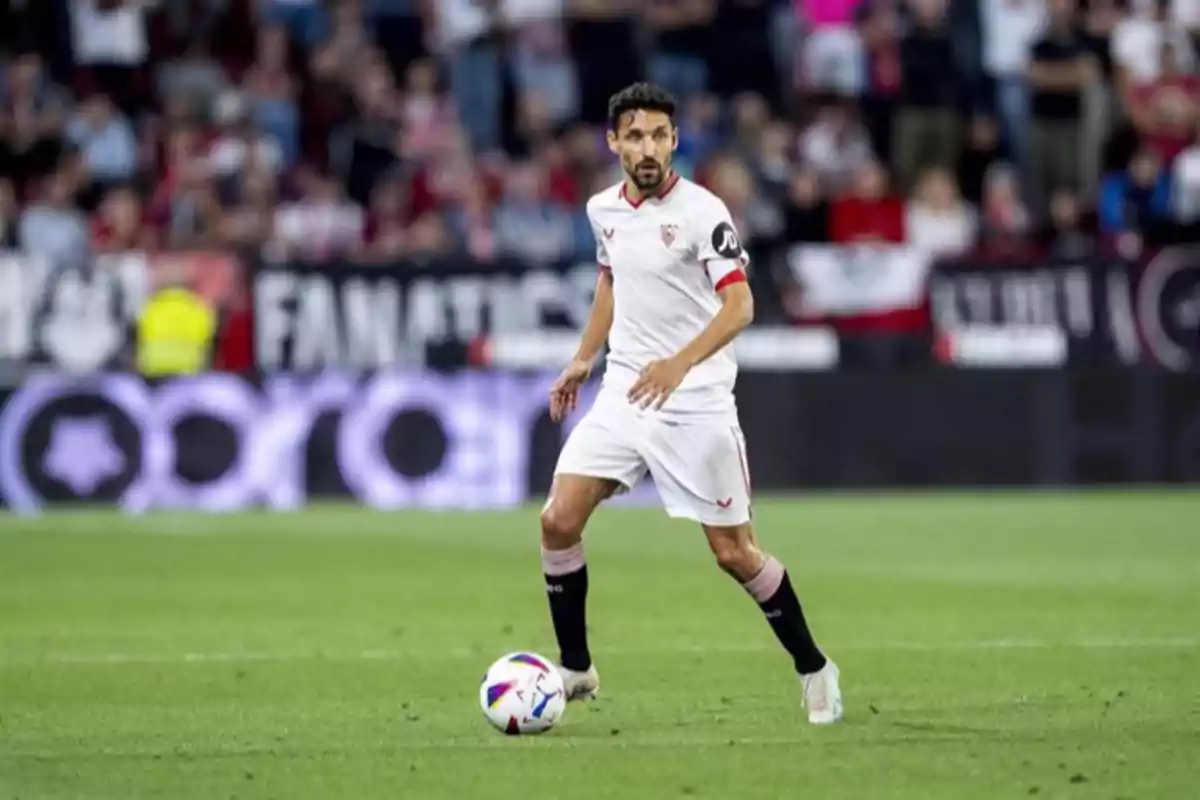 Un jugador de fútbol con uniforme blanco y detalles rojos controla el balón en un campo de juego con espectadores de fondo.