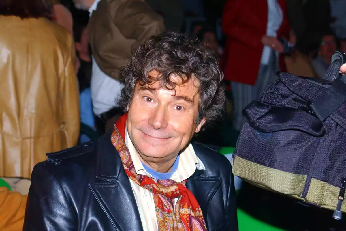 Un hombre sonriente con cabello rizado y una bufanda colorida, vistiendo una chaqueta de cuero, rodeado de personas en un evento.